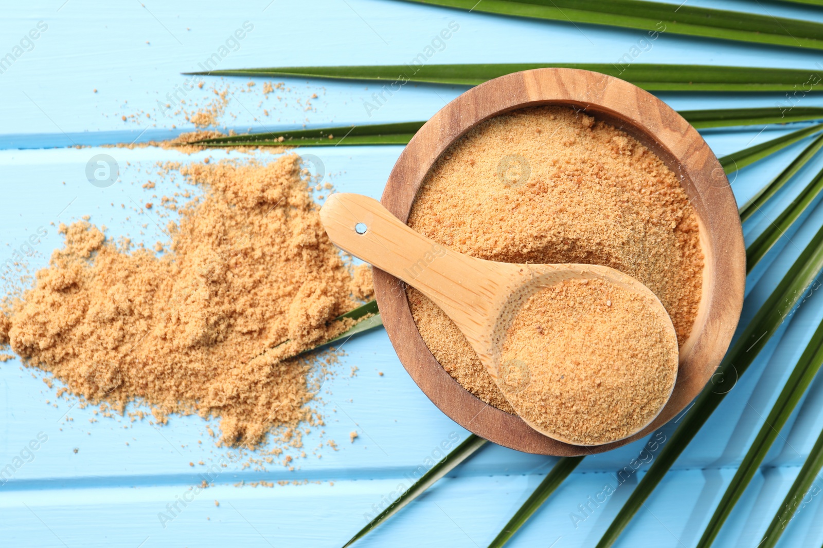 Photo of Coconut sugar in bowl, spoon and palm leaves on light blue wooden table, flat lay