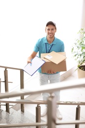 Photo of Young courier standing with parcels and clipboard on stairs indoors