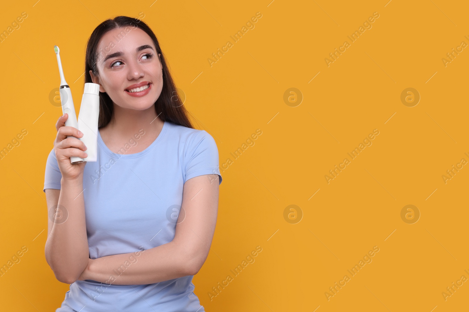 Photo of Happy young woman holding electric toothbrush and tube of toothpaste on yellow background, space for text