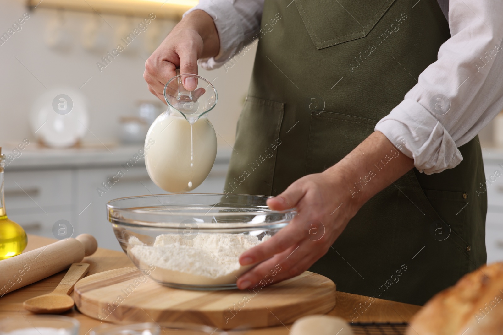 Photo of Making bread. Man pouring milk into bowl with flour at wooden table in kitchen, closeup