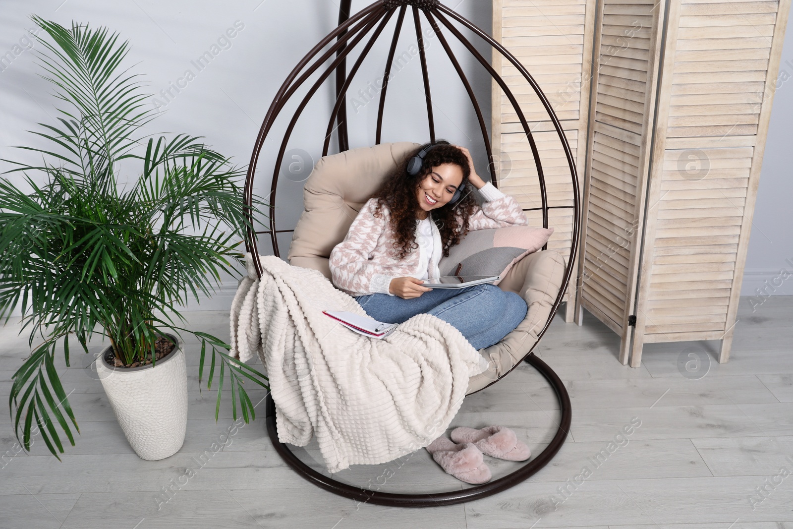 Photo of African American woman with headphones and tablet studying in egg chair at home. Distance learning