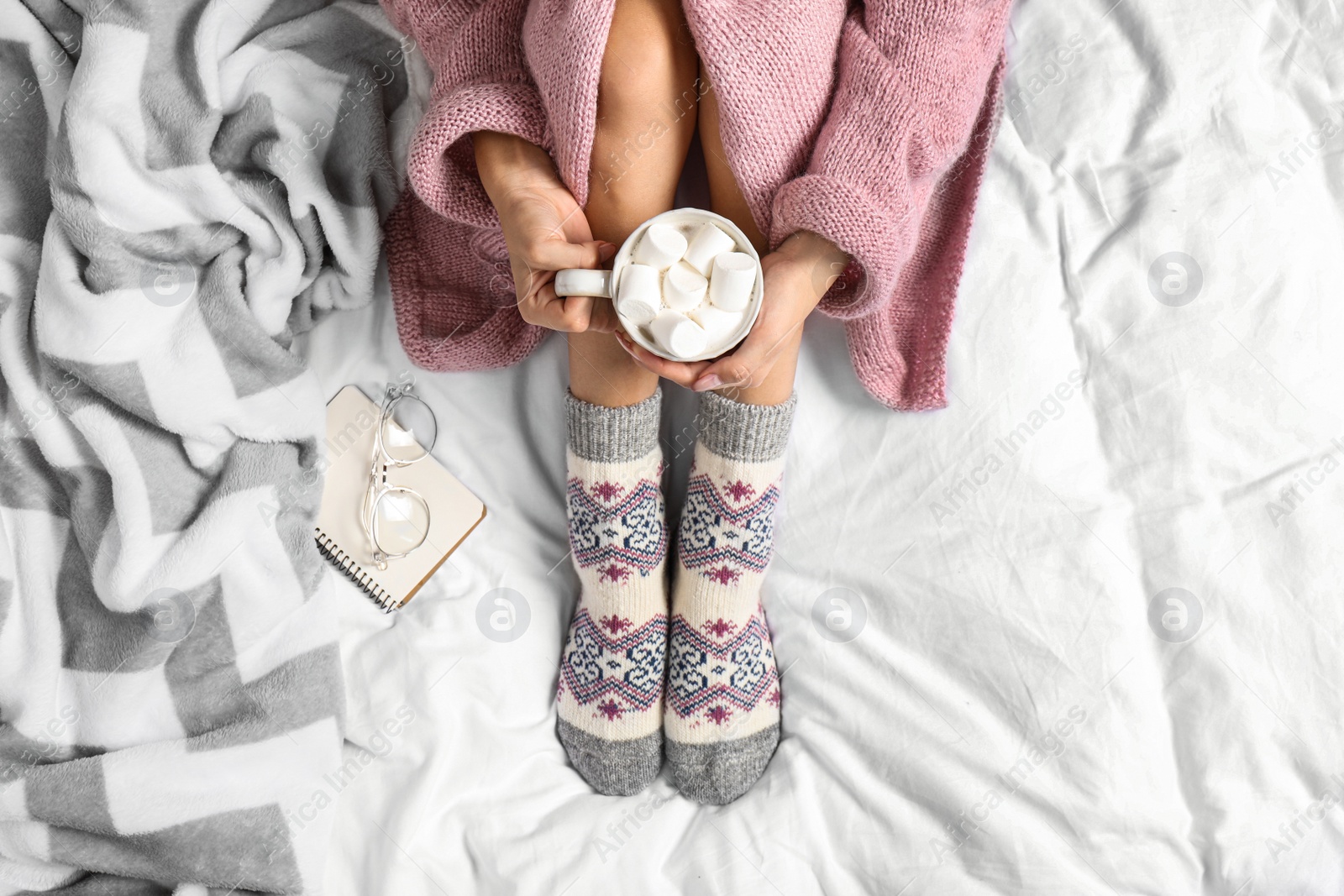 Photo of Woman with cocoa wearing knitted socks on white fabric, top view. warm clothes