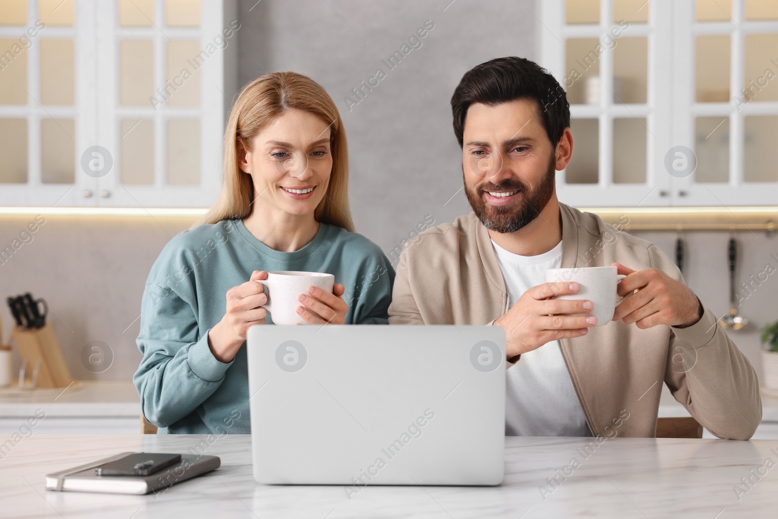 Photo of Happy couple with laptop and cups of drink at white table in kitchen