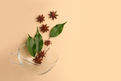 Anise stars and green leaves falling into glass cup on beige background, flat lay. Space for text