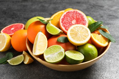 Photo of Different fresh citrus fruits and leaves in bowl on grey textured table, closeup