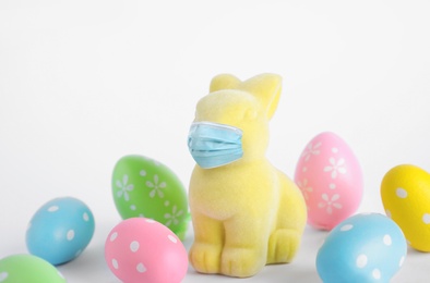 Image of COVID-19 pandemic. Easter bunny toy in protective mask and dyed eggs on white background