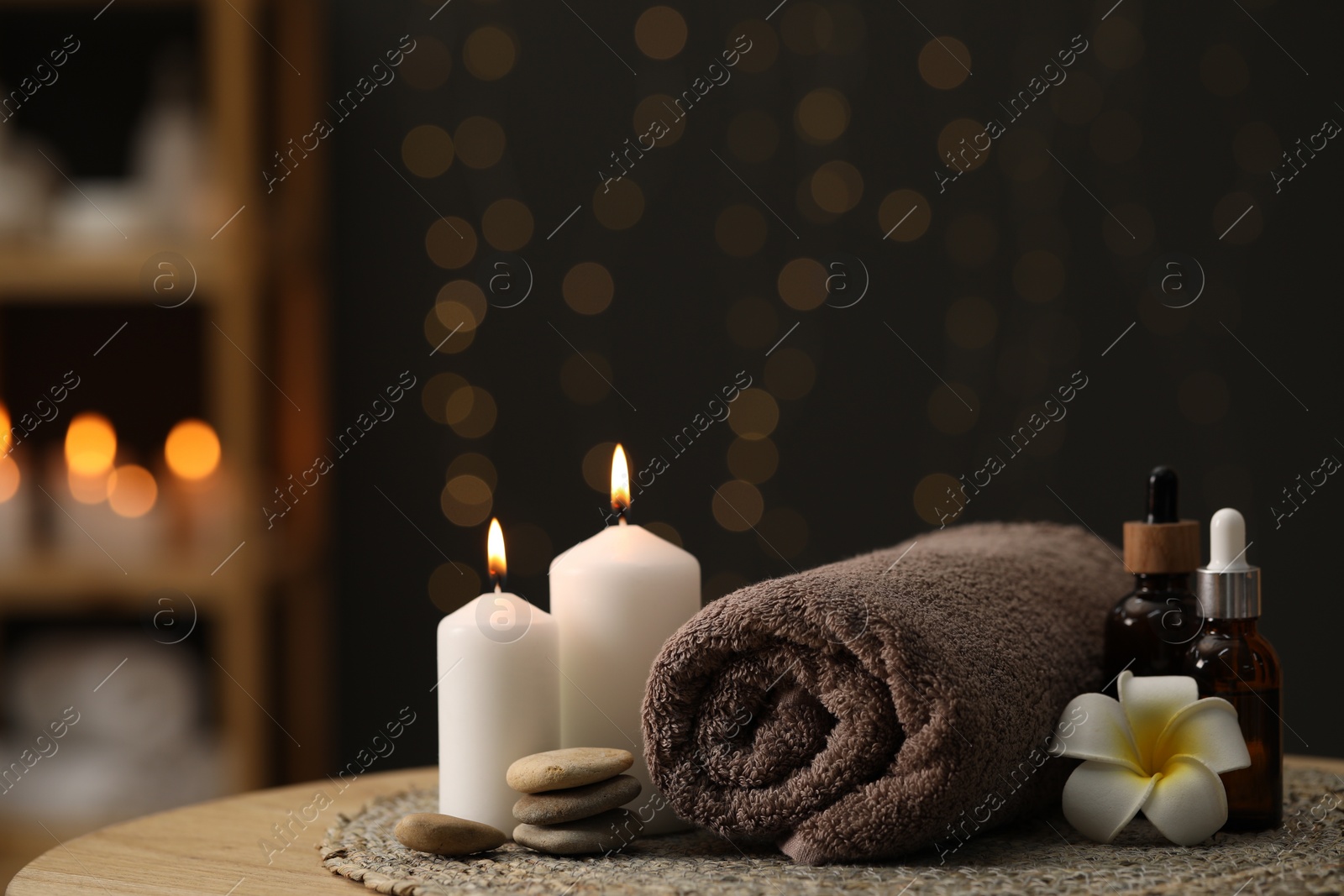 Photo of Spa composition. Rolled towel, cosmetic products, stones, burning candles and plumeria flower on table indoors. Space for text