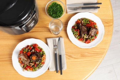 Photo of Plates with meat and garnish prepared in multi cooker on wooden table, flat lay