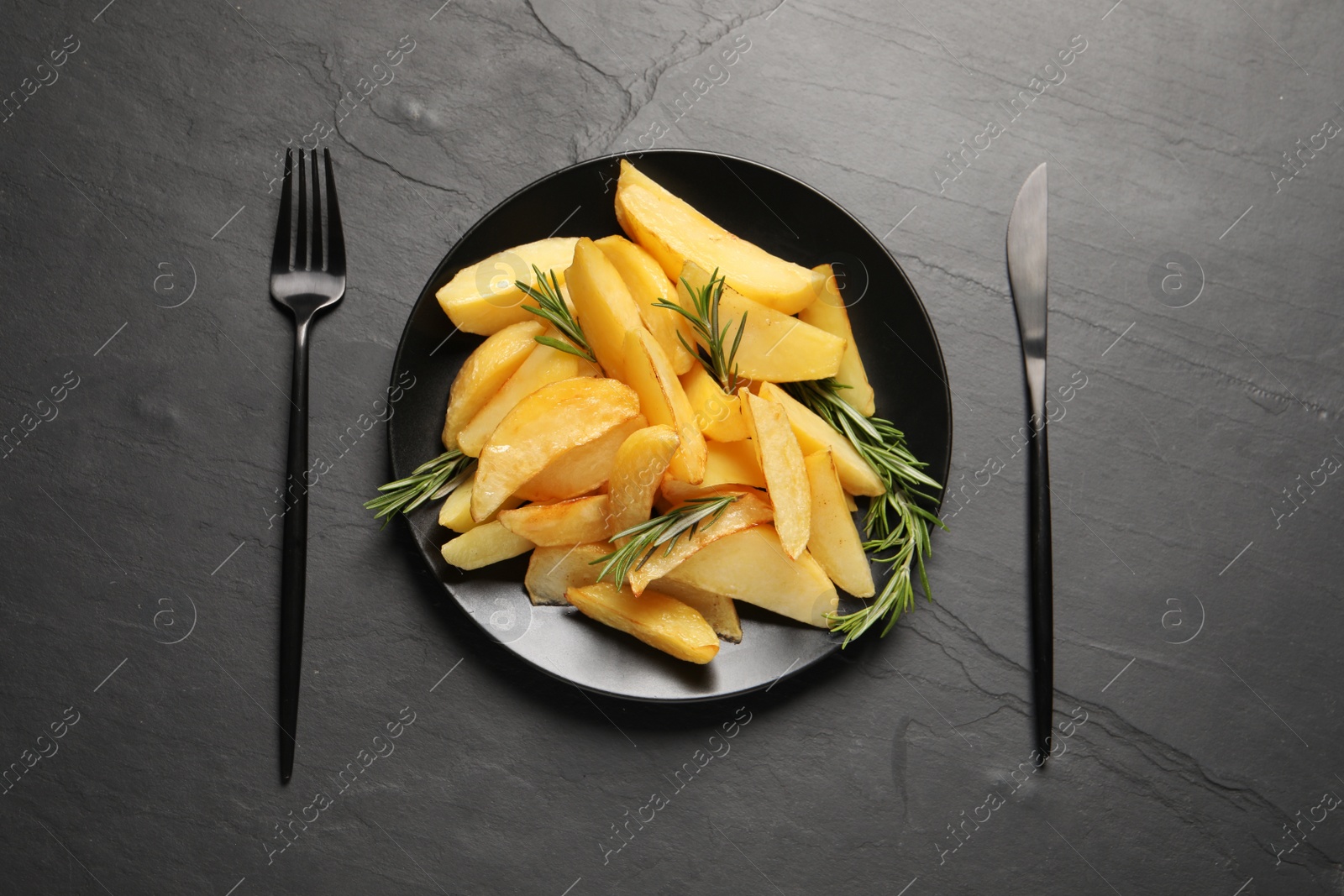 Photo of Served tasty baked potato wedges and rosemary on black table, flat lay