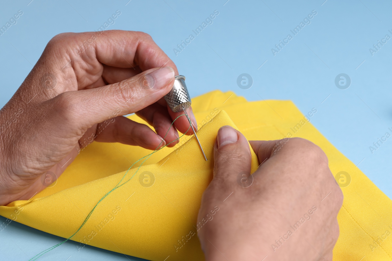 Photo of Woman sewing on yellow cloth with thimble and needle against light blue background, closeup