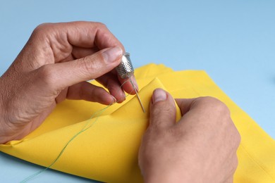 Photo of Woman sewing on yellow cloth with thimble and needle against light blue background, closeup