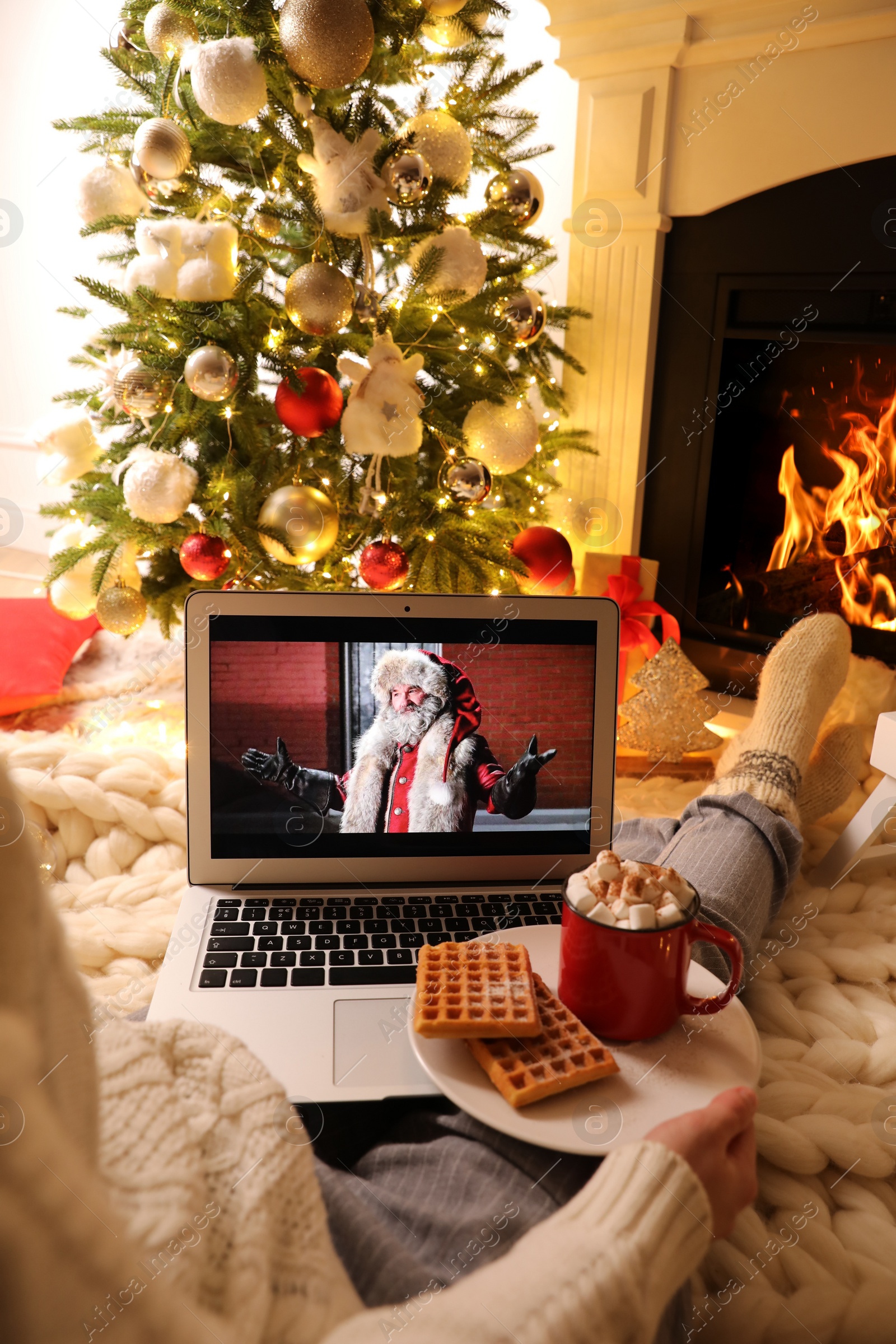 Photo of MYKOLAIV, UKRAINE - DECEMBER 23, 2020: Woman with cocoa and wafers watching The Christmas Chronicles movie on laptop near fireplace at home, closeup. Cozy winter holidays atmosphere