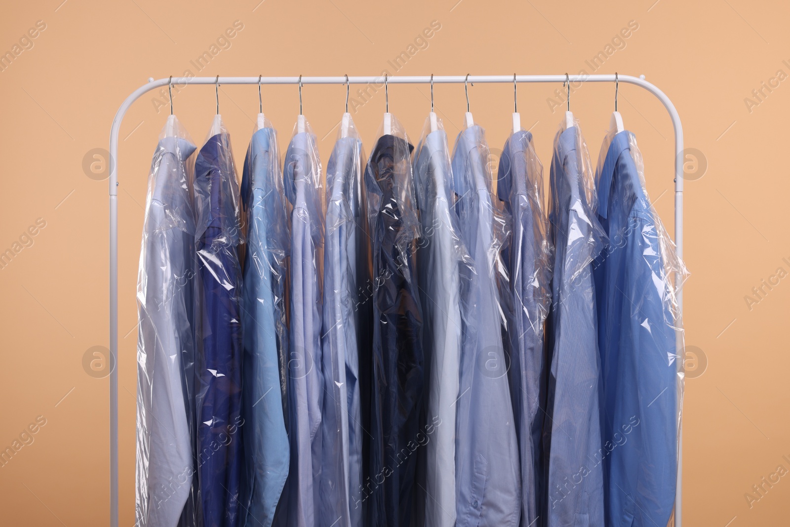 Photo of Dry-cleaning service. Many different clothes in plastic bags hanging on rack against beige background