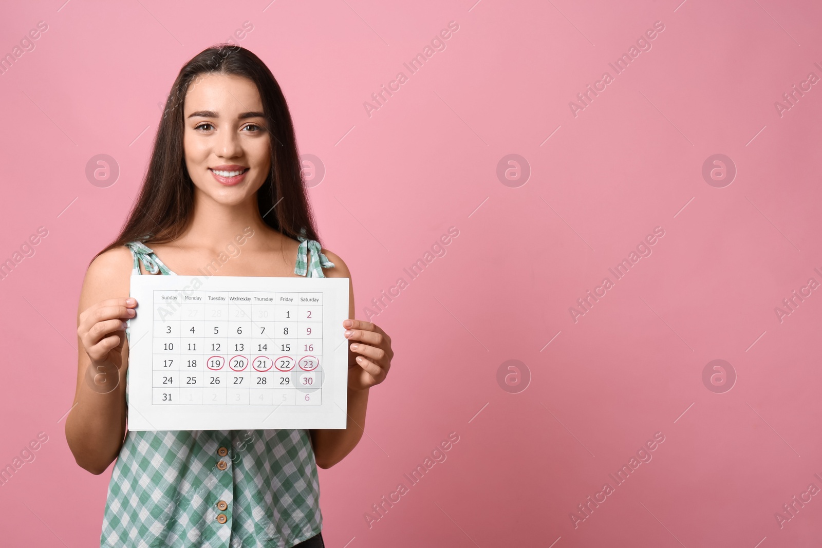 Photo of Young woman holding calendar with marked menstrual cycle days on pink background. Space for text