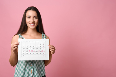 Young woman holding calendar with marked menstrual cycle days on pink background. Space for text