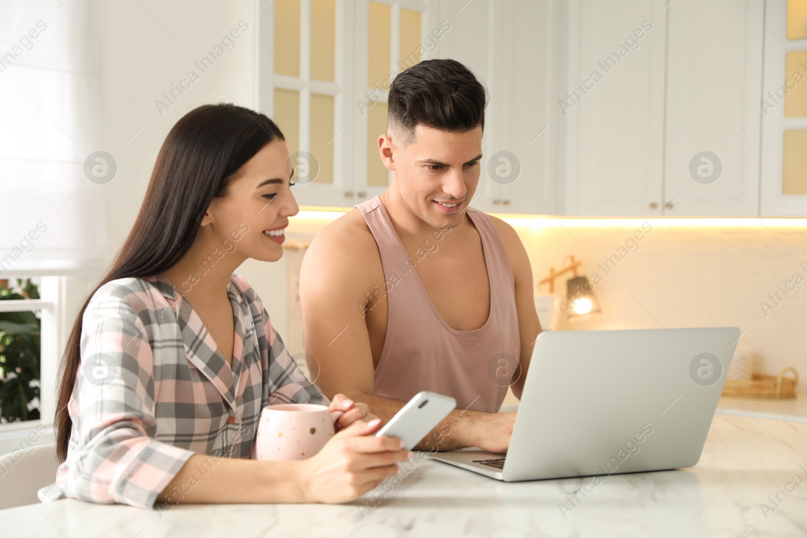 Photo of Happy couple wearing pyjamas with gadgets spending time together in kitchen