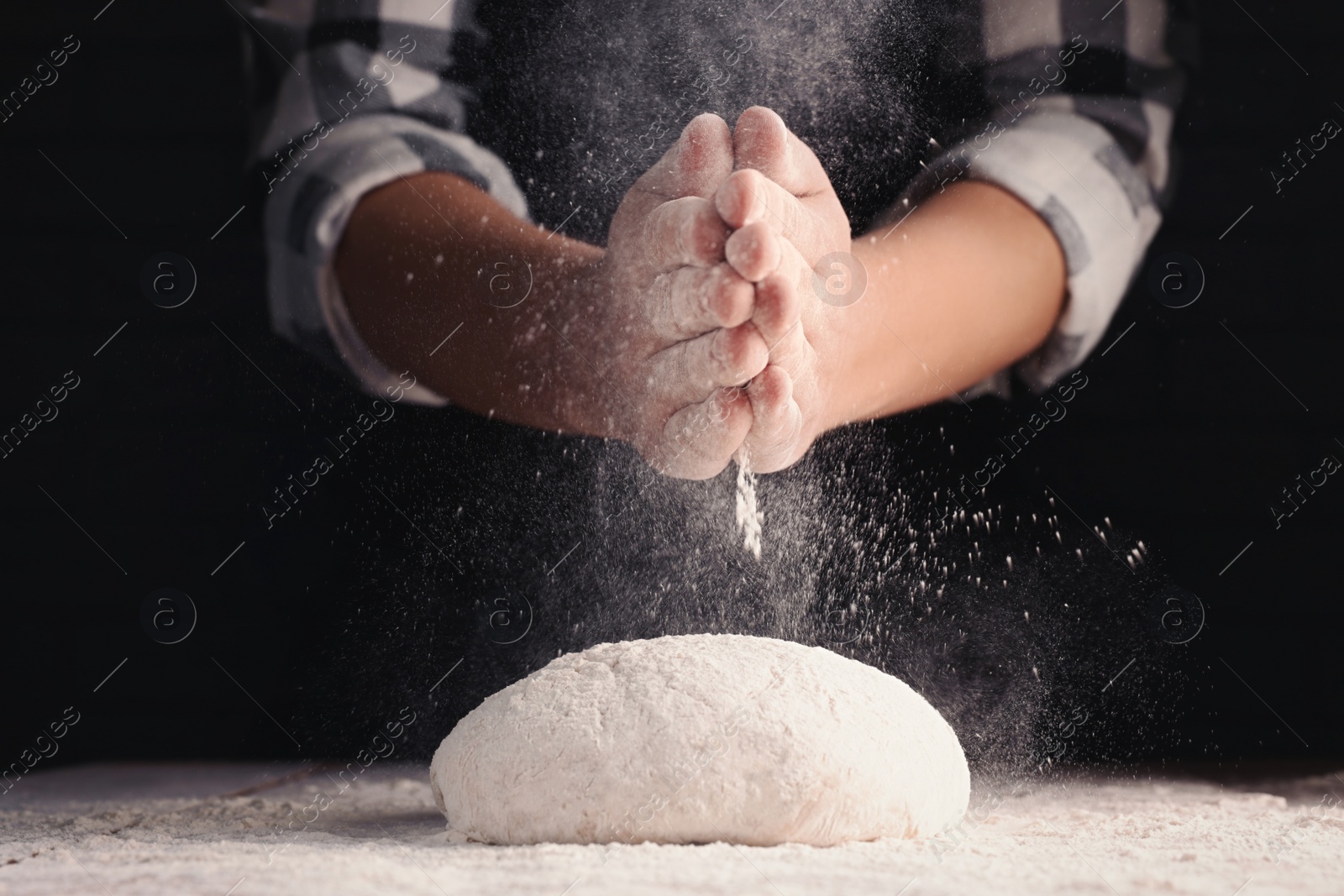 Photo of Man sprinkling flour over dough at wooden table on dark background, closeup