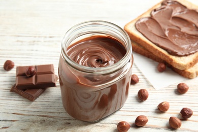 Photo of Glass jar with tasty chocolate cream, bread and hazelnuts on wooden table