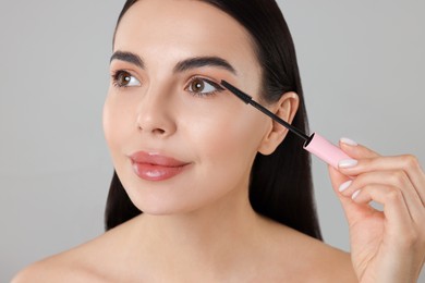 Beautiful young woman applying mascara on grey background, closeup