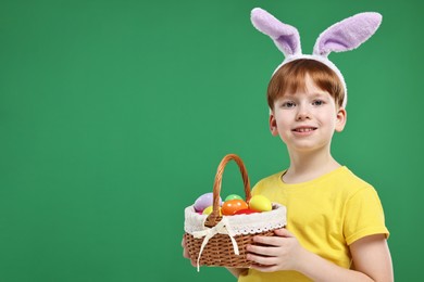 Easter celebration. Cute little boy with bunny ears and wicker basket full of painted eggs on green background. Space for text