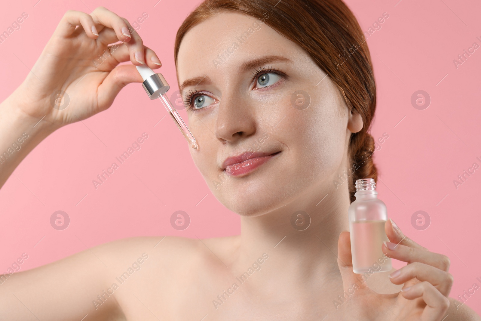 Photo of Beautiful woman with freckles applying cosmetic serum onto her face on pink background, closeup