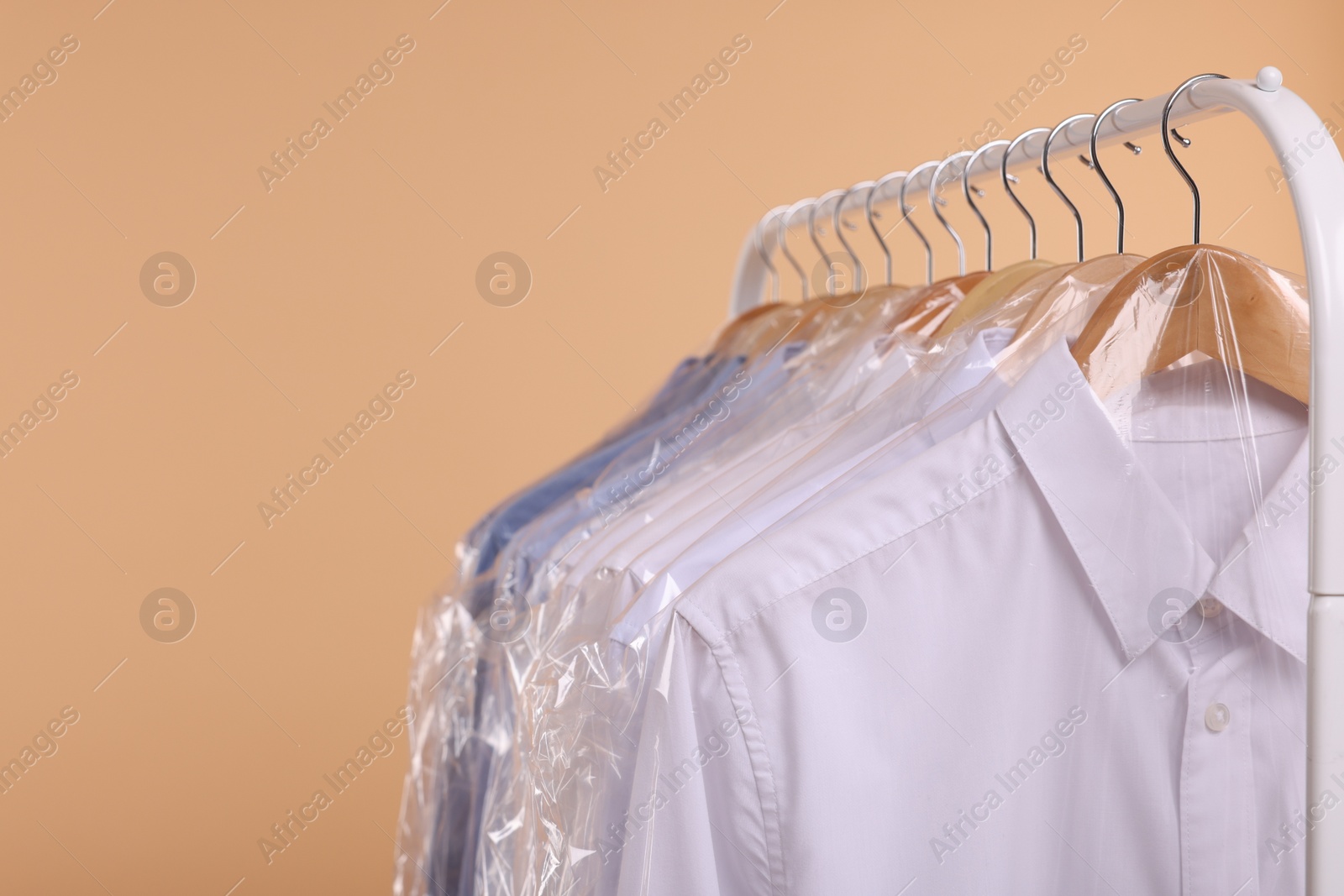 Photo of Dry-cleaning service. Many different clothes in plastic bags hanging on rack against beige background, closeup and space for text