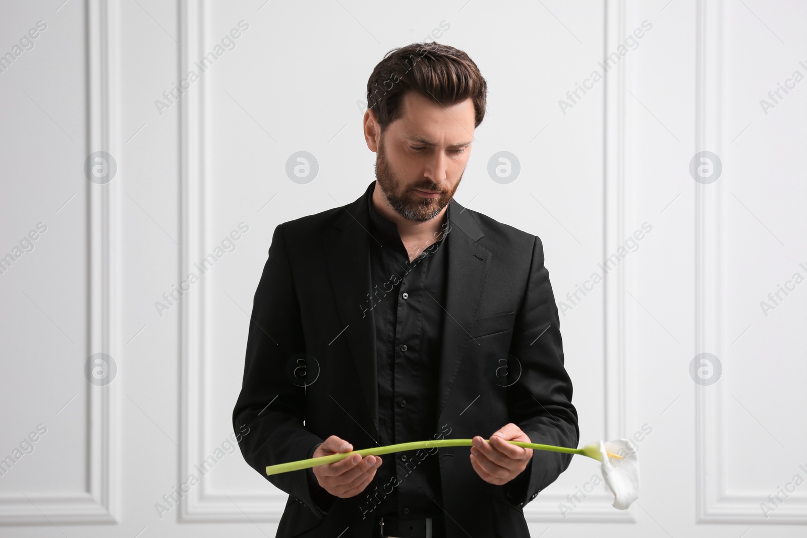 Photo of Sad man with calla lily flower near white wall. Funeral ceremony