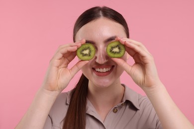 Smiling woman covering eyes with halves of kiwi on pink background