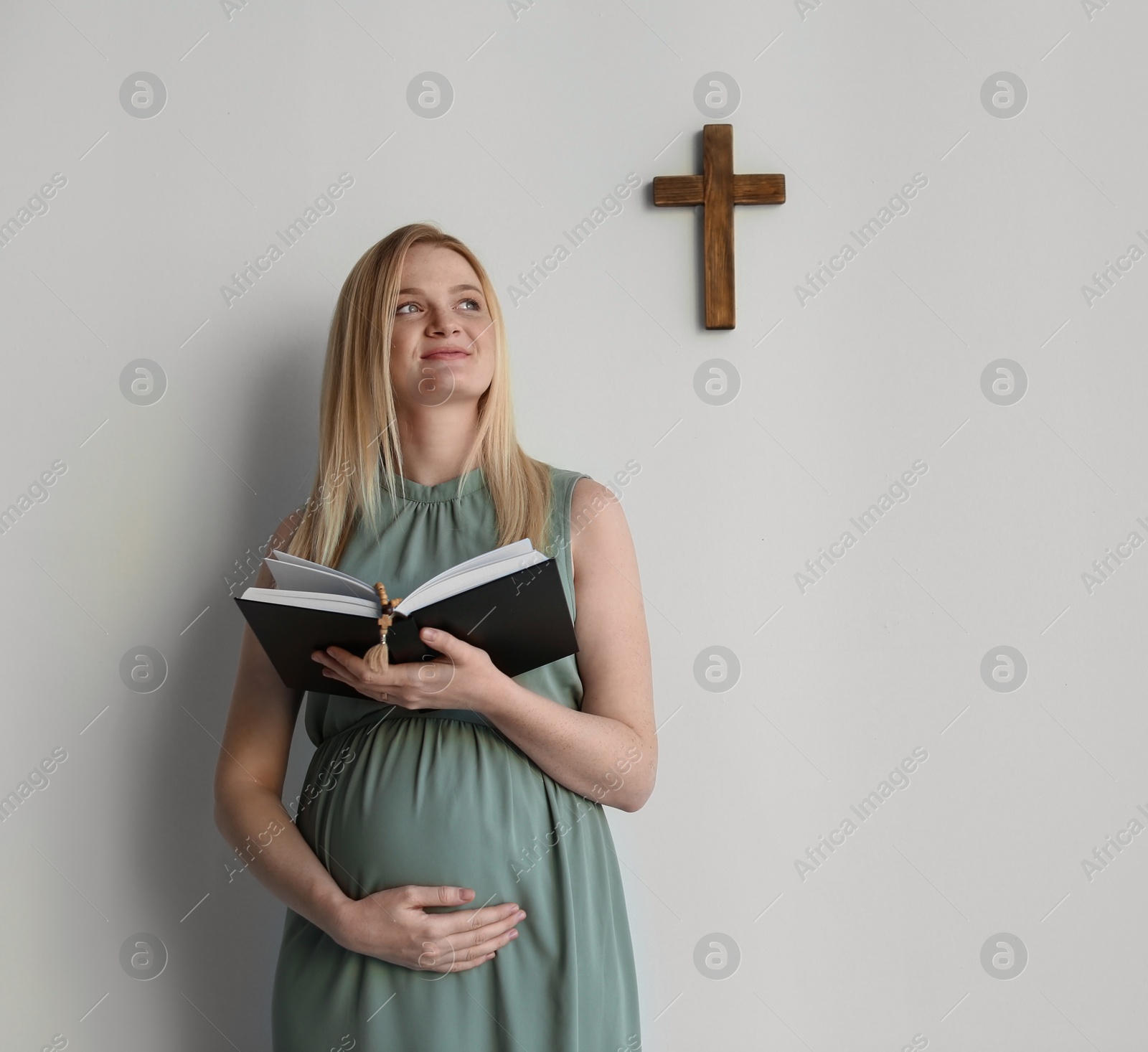 Photo of Young pregnant woman with Bible praying near light wall. Space for text