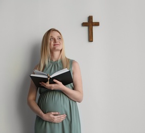 Young pregnant woman with Bible praying near light wall. Space for text