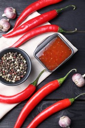 Spicy chili sauce, garlic, peppers and peppercorns on black wooden table, flat lay