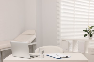 Photo of Doctor's workplace. Laptop, clipboard and glass of water on white table in hospital