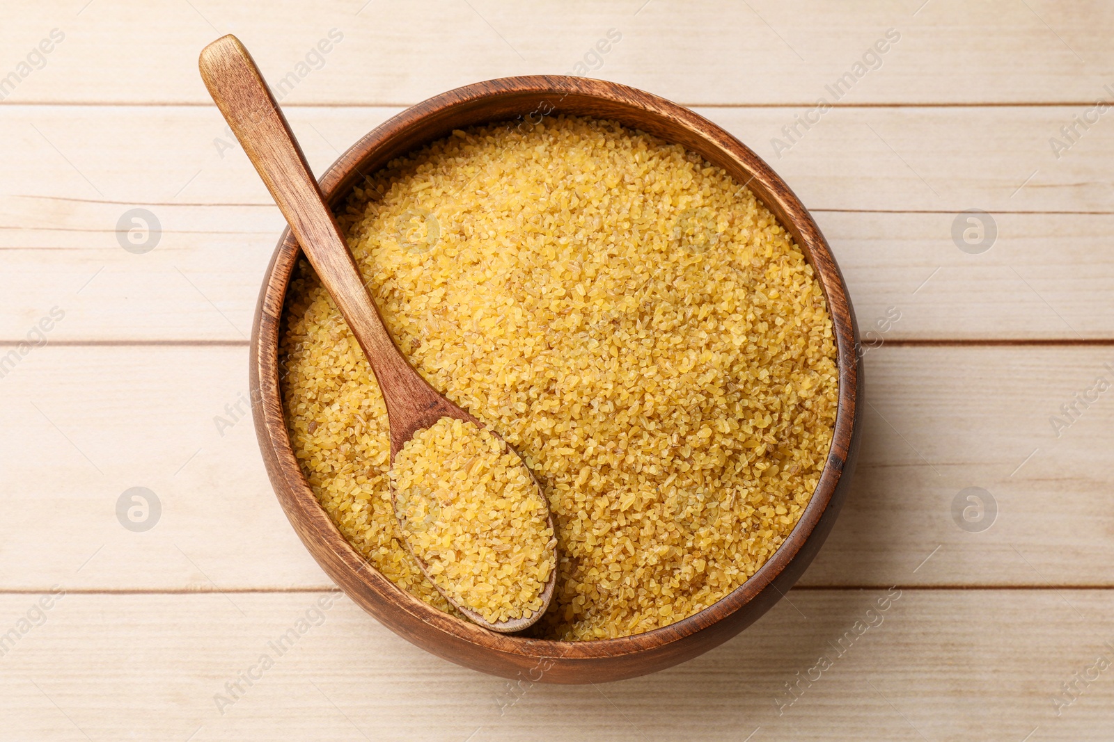 Photo of Bowl and spoon with raw bulgur on light wooden table, top view