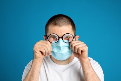 Photo of Man wiping foggy glasses caused by wearing disposable mask on blue background. Protective measure during coronavirus pandemic