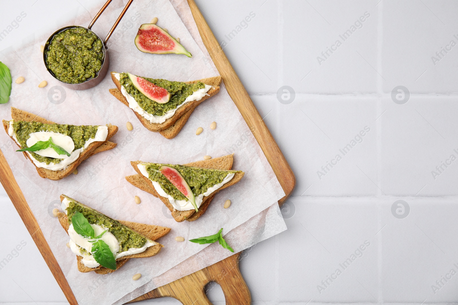 Photo of Tasty bruschettas with cream cheese, pesto sauce, figs and fresh basil on white tiled table, top view. Space for text