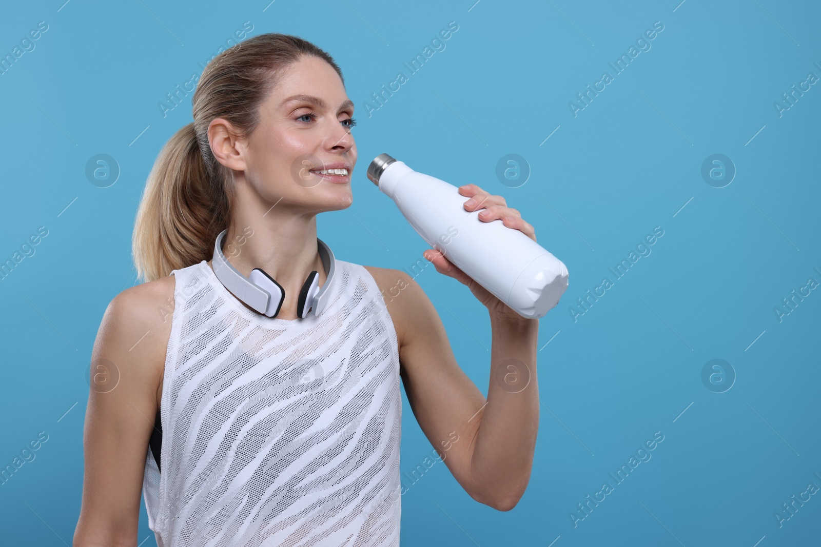 Photo of Sportswoman with thermo bottle and headphones on light blue background, space for text