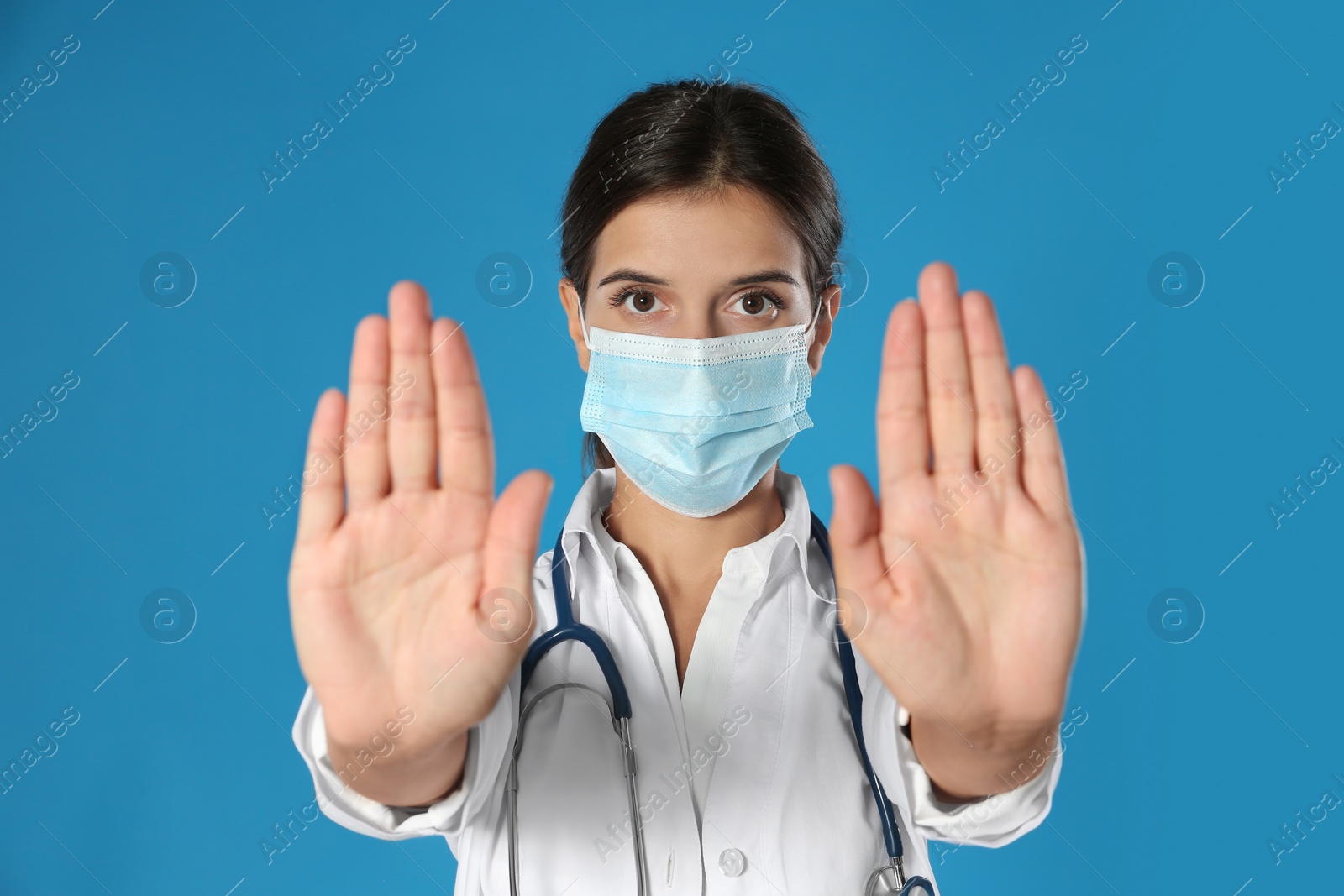 Photo of Doctor in protective mask showing stop gesture on blue background. Prevent spreading of coronavirus