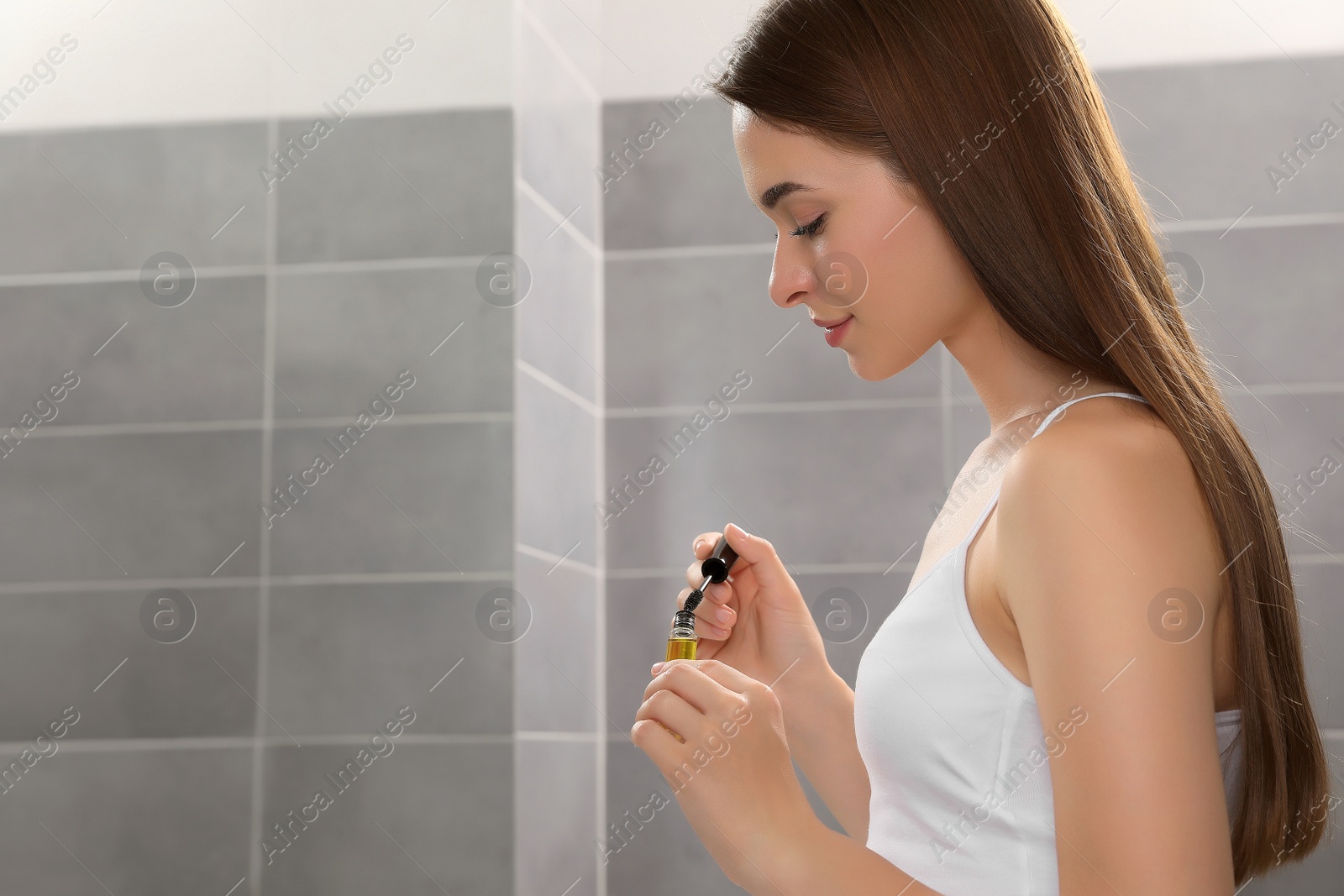 Photo of Young woman with eyelash oil in bathroom, space for text