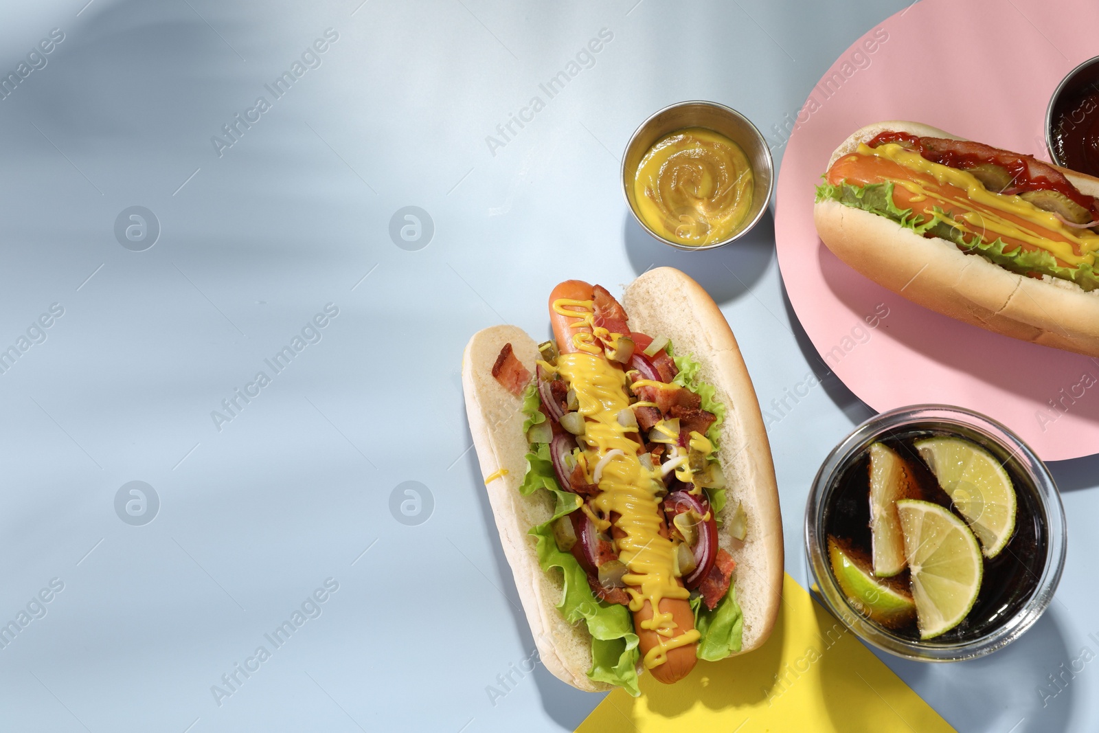 Photo of Delicious hot dogs with bacon, lettuce, onion and pickle served on light blue table, flat lay. Space for text