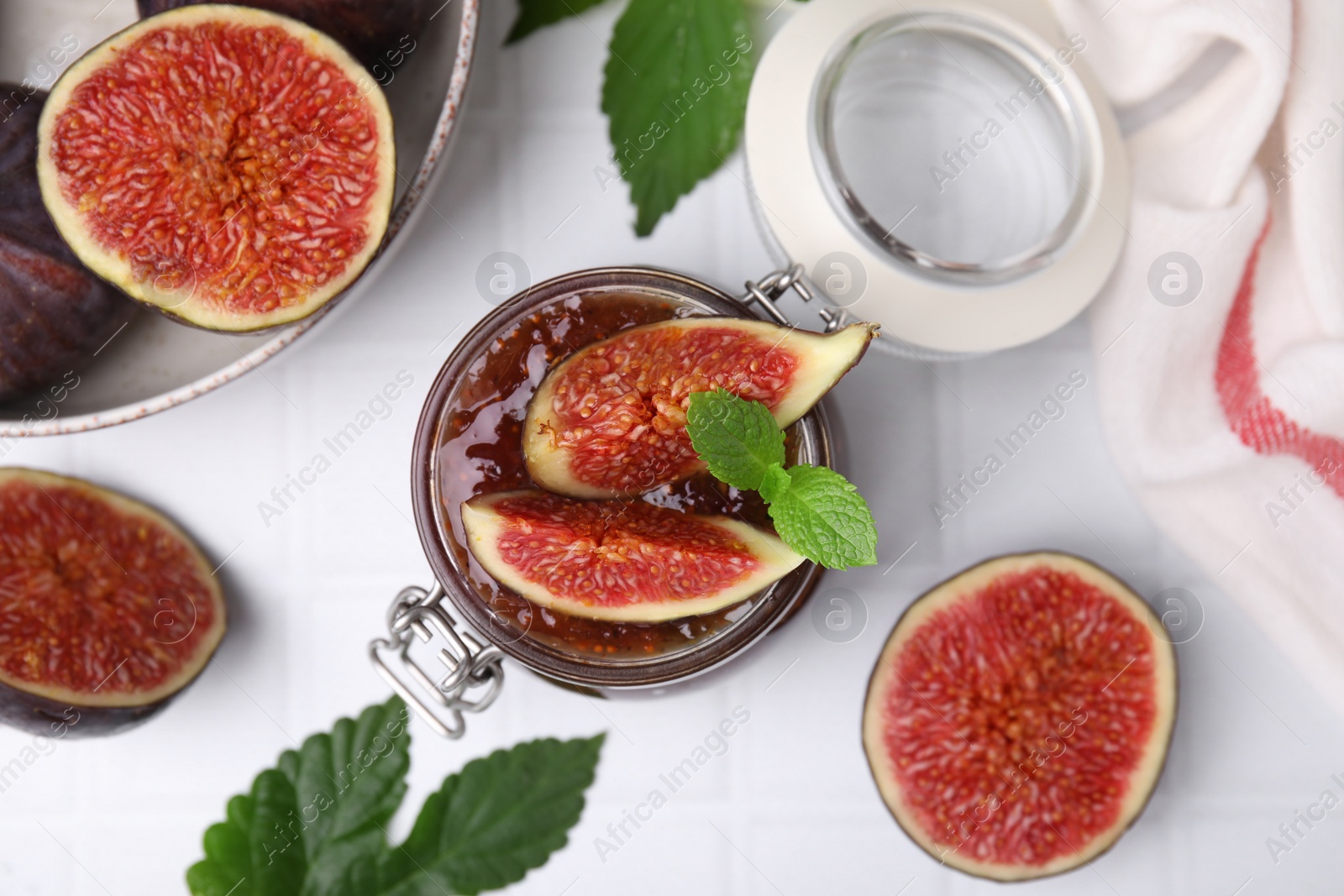 Photo of Glass jar of tasty sweet fig jam and fruits on white tiled table, flat lay