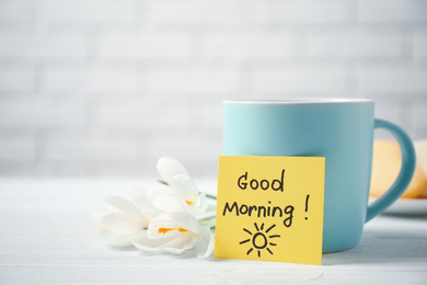 Photo of Delicious coffee, flowers and card with GOOD MORNING wish on white wooden table, closeup. Space for text