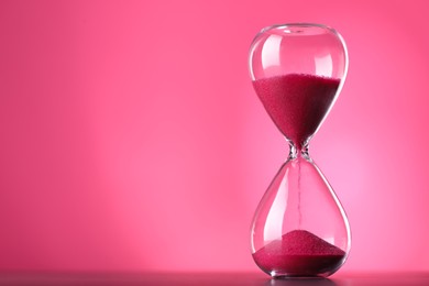 Photo of Hourglass with flowing sand on table against pink background, space for text