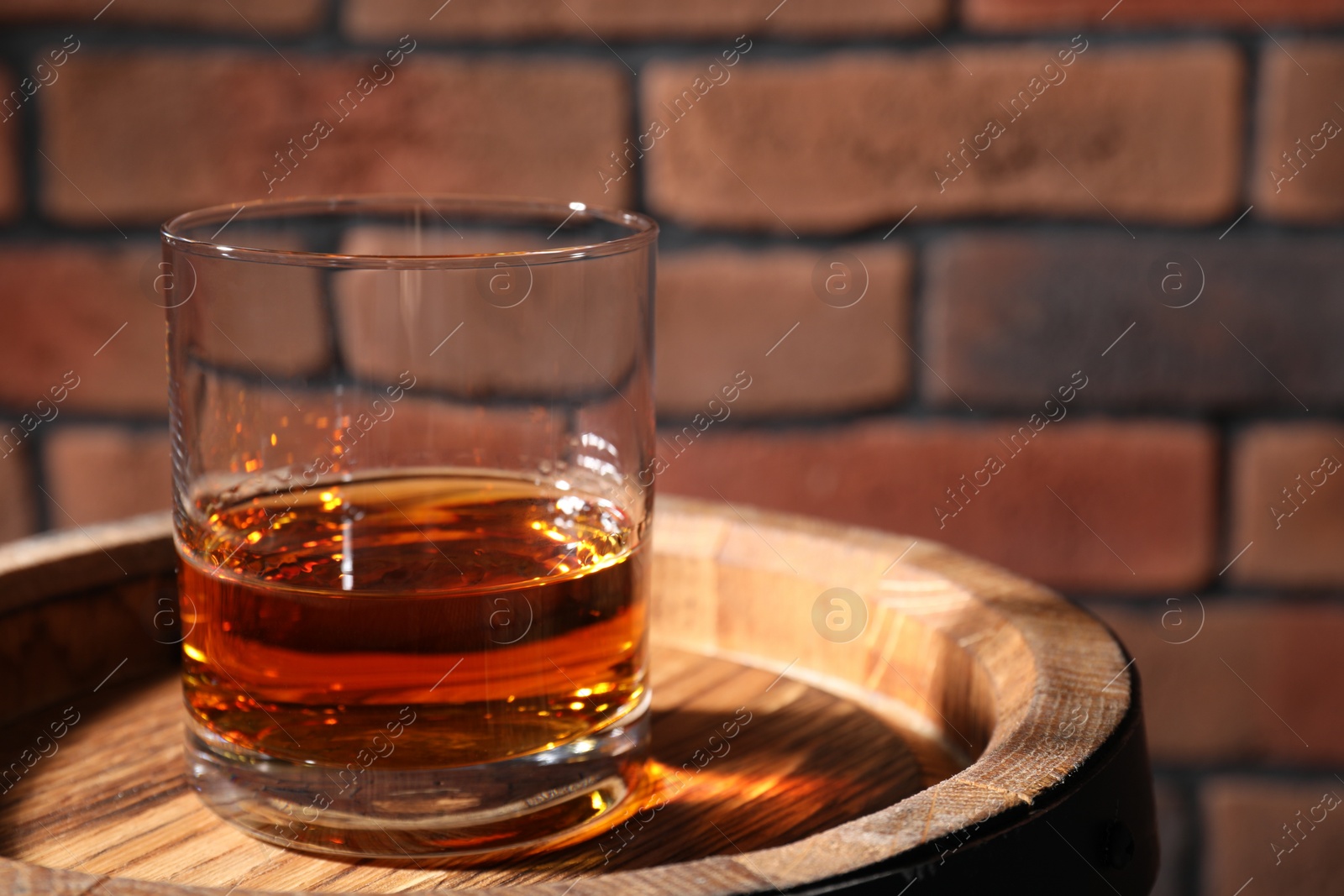 Photo of Glass of whiskey on wooden barrel against brick wall, closeup. Space for text