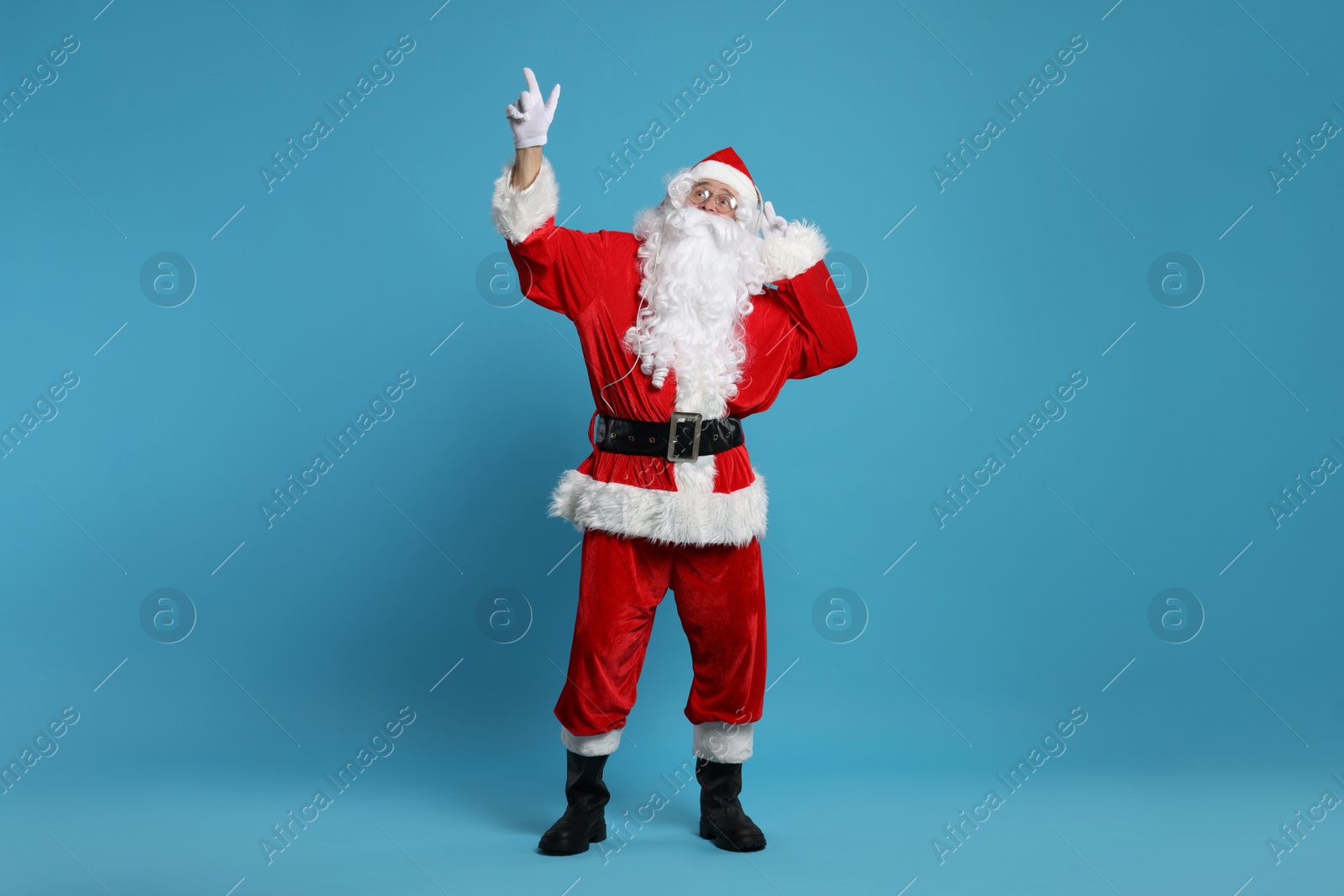 Photo of Merry Christmas. Santa Claus in headphones listening to music and pointing at something on light blue background