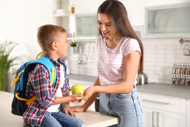 Happy mother giving apple to little child's with school bag in kitchen