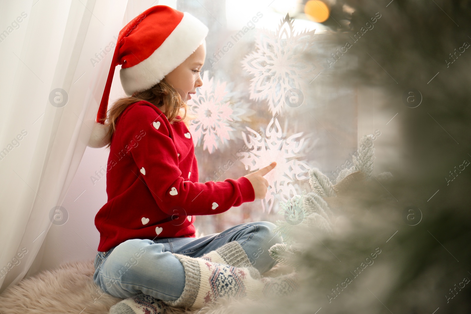 Photo of Little girl in Santa hat near window decorated with paper snowflakes at home