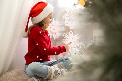 Photo of Little girl in Santa hat near window decorated with paper snowflakes at home