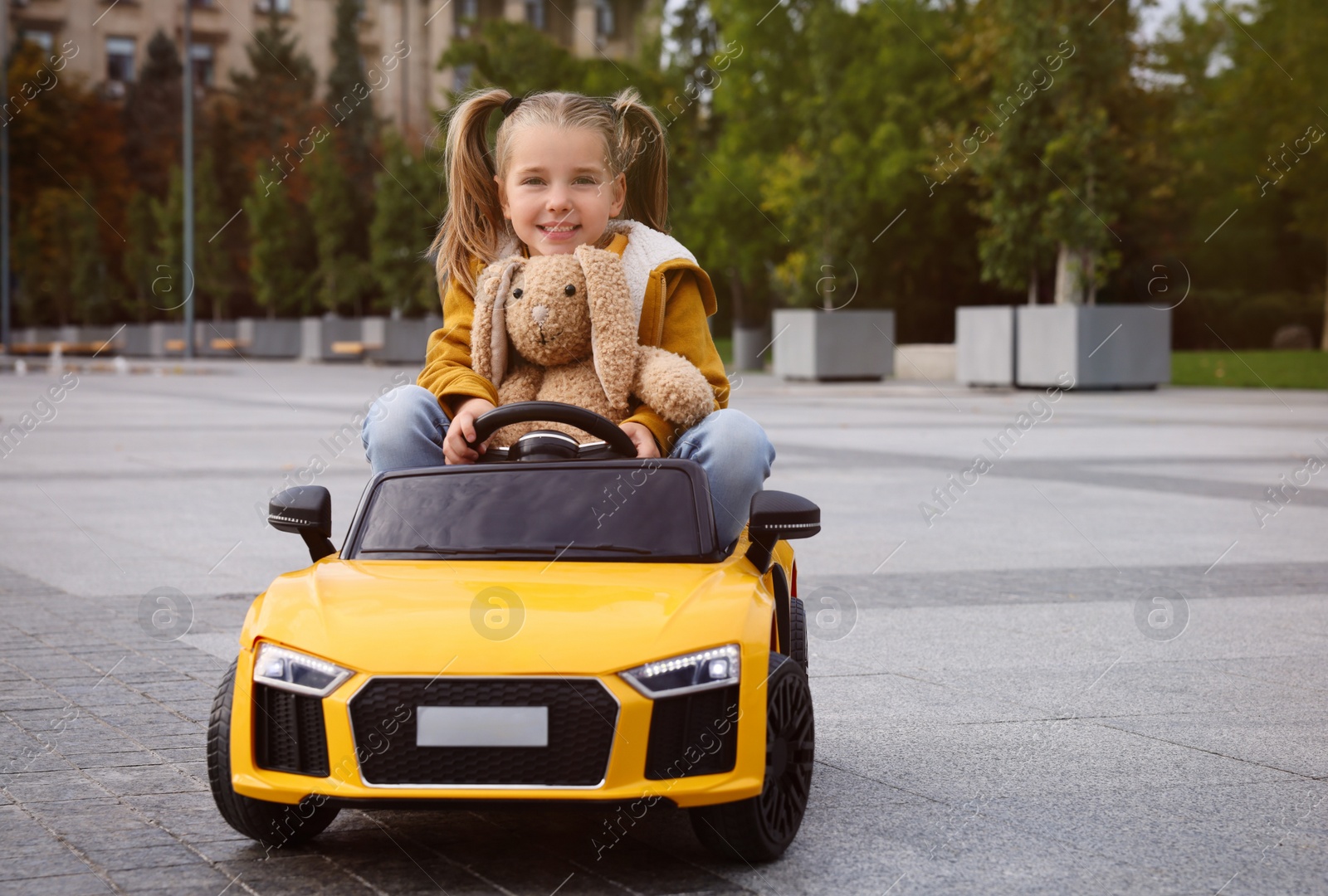 Photo of Cute little girl with toy bunny driving children's car on city street. Space for text
