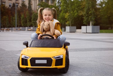 Photo of Cute little girl with toy bunny driving children's car on city street. Space for text