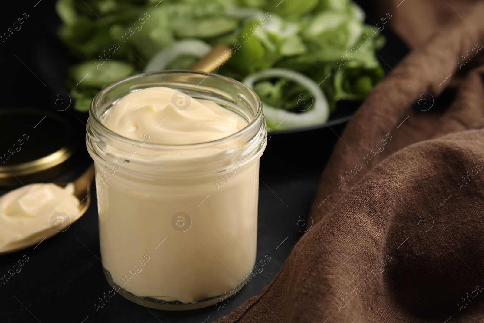 Photo of Jar and spoon with delicious mayonnaise near fresh salad on black table